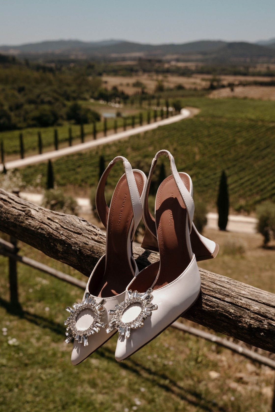 Crystal-embellished slingback bridal heels on a wooden fence rail, Tuscan vineyard and cypress trees behind them