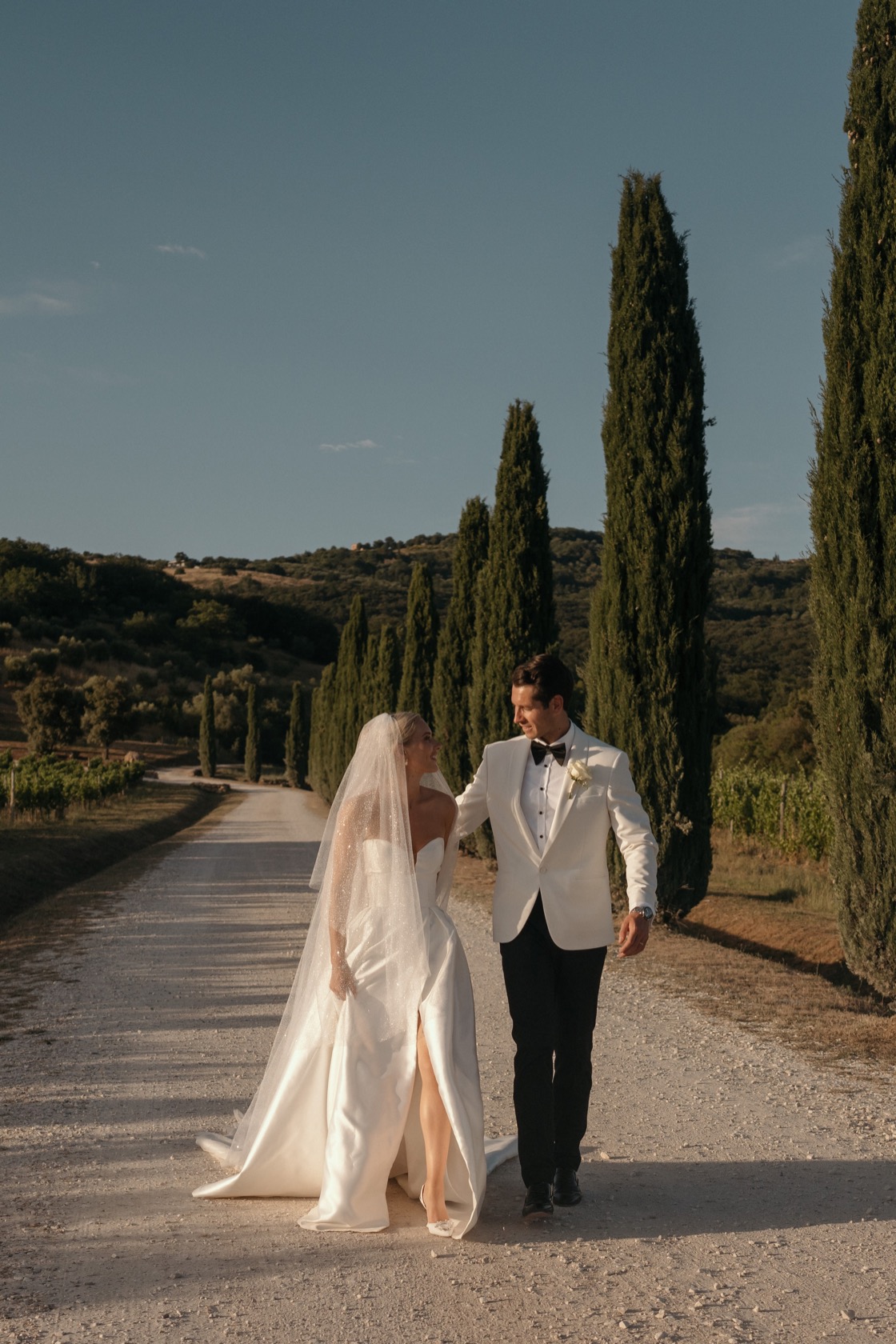 Bride in strapless white gown and cathedral veil and groom in white dinner jacket walking hand in hand along a cypress-lined drive at Conti di San Bonifacio Wine Resort, Tuscany.