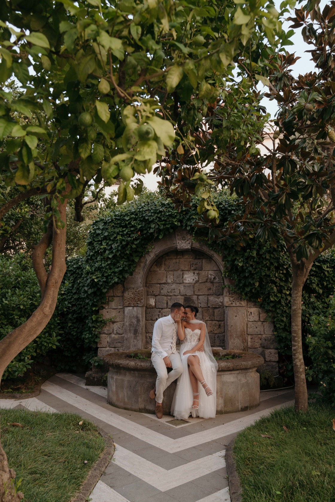 Jade in a strapless tulle gown and Matt in a white suit sitting together at an ivy-covered stone fountain in the garden courtyard, surrounded by lemon trees at The Cloisters