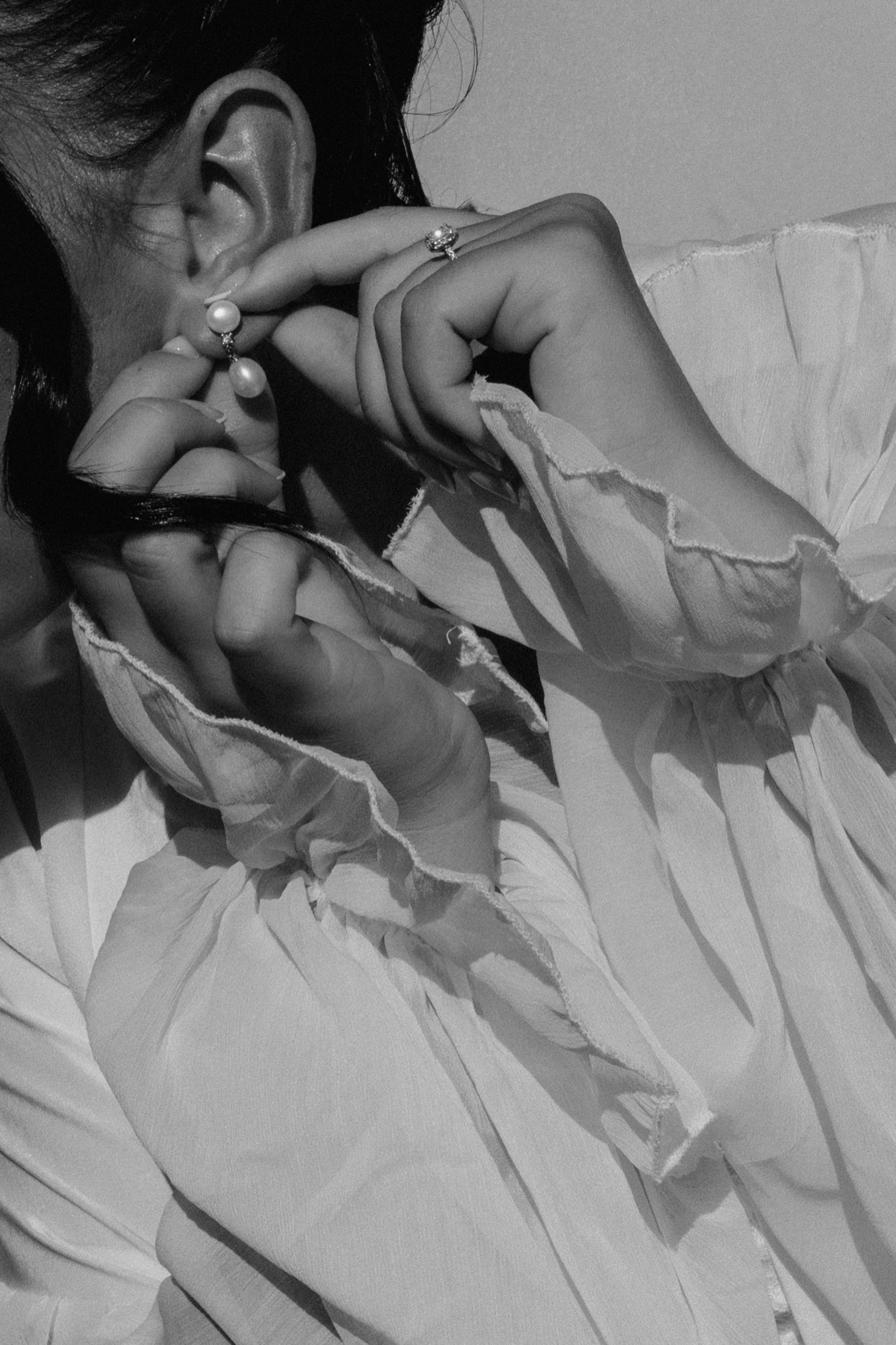 Close-up black and white photograph of Jade fastening a pearl drop earring, diamond ring visible, ruffled white bridal gown sleeve in bright natural light