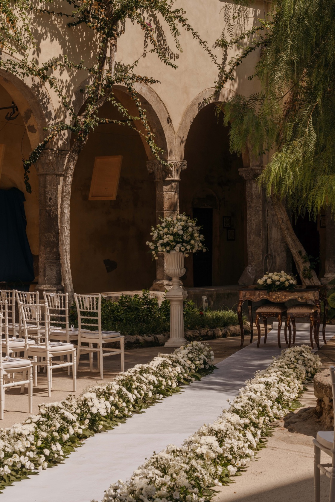 White floral carpet lining the ceremony aisle inside the stone-arched cloister courtyard, white chiavari chairs and a tall urn of white roses in dappled sunlight