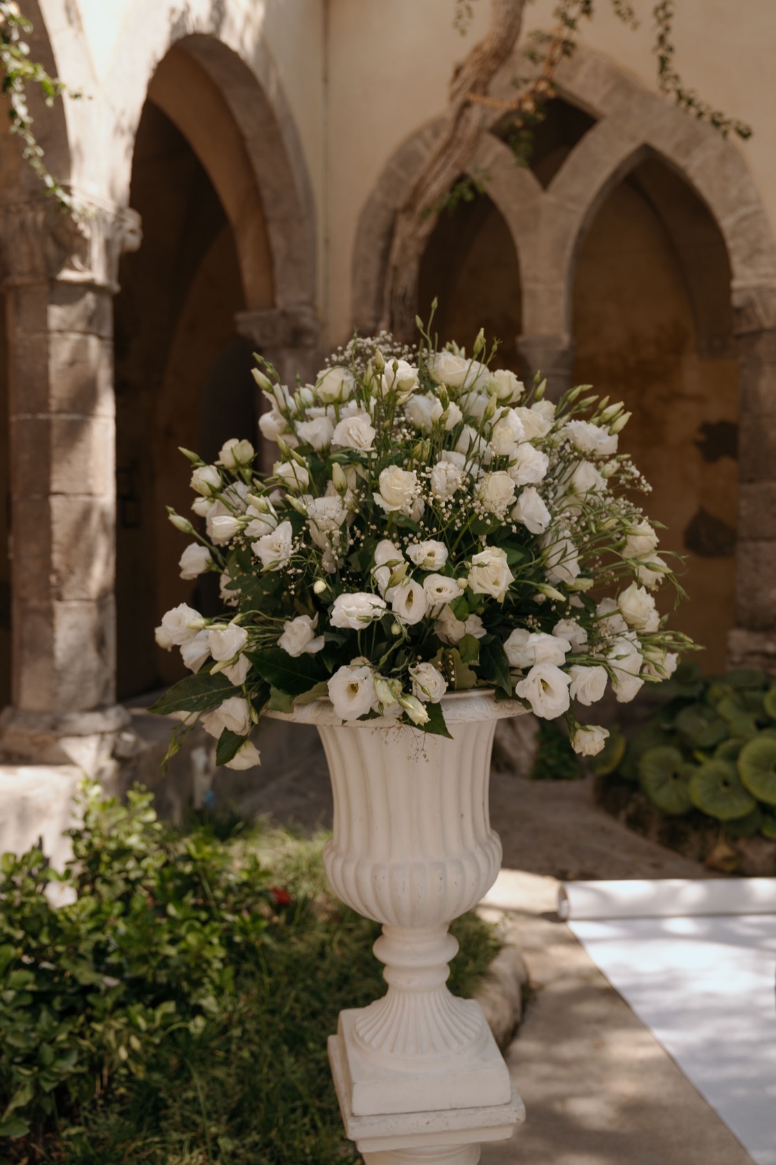 Large white floral urn of lisianthus, gypsophila and greenery on a stone pedestal beside the white aisle, Gothic stone arches softly blurred behind