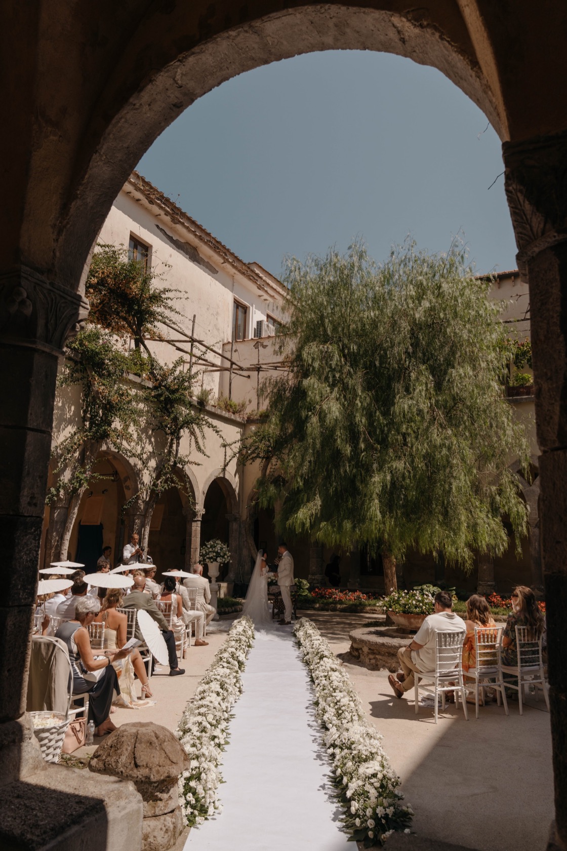 Wedding ceremony in progress at The Cloisters viewed through a wide stone arch — white aisle runner, low white floral arrangements, chiavari chairs, weeping willow tree, deep blue sky above