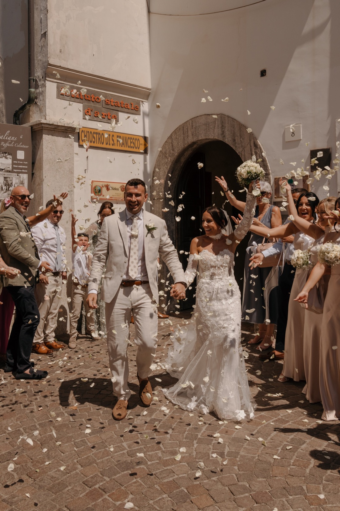 Jade in lace gown and Matt in ivory suit walk through a shower of white rose petals thrown by guests on the sunlit cobblestone courtyard outside a stone archway